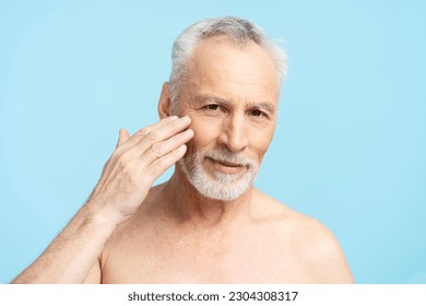 Portrait of handsome happy gray haired senior man applying anti aging face cream looking in mirror isolated on blue background. Morning routine, skin care, beauty procedures concept - Powered by Shutterstock