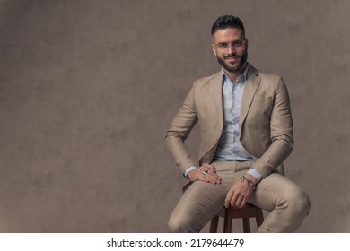 Portrait Of Handsome Happy Guy In Suit With Open Collar Shirt Smiling While Sitting On Wooden Chair In Front Of Beige Background 