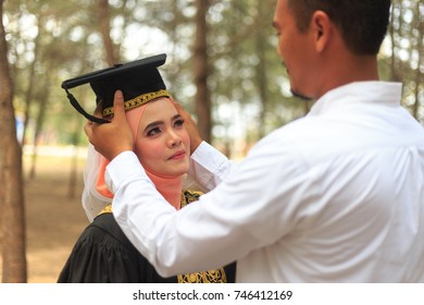 Portrait Of Handsome And Helpful Guy Helping A Needy Young Woman Wearing Her Graduation Hat Or Mortarboard. Lovely Couple, Selective Focus In Nature Background