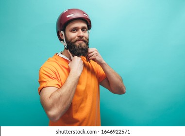 Portrait Of Handsome Hipster Guy With Beard Wearing Blank Orange T-shirt And Red Bike Helmet , Isolated On Blue Studio Wall.