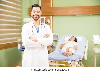 Portrait Of A Handsome Hispanic Obgyn Doctor Standing In A Hospital Room With A Patient And Smiling