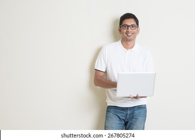 Portrait Of Handsome Indian Guy Using Laptop Computer, Standing On Plain Background With Shadow, Copy Space At Side.