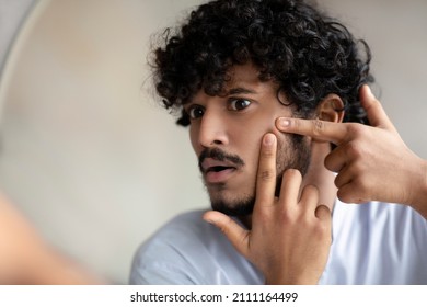 Portrait of handsome indian man touching his face, pressing pimple on face with fingers, looking at mirror, closeup. Healthcare, stressful lifestyle hygienic acne concept