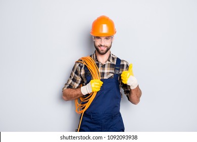 Portrait of handsome joyful electrician in hardhat, overall, shirt with bristle, holding rolled wires on shoulder, showing thumb up recommend approve sign over grey background
