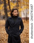 Portrait of handsome long haired man in black trench coat in the park in wonderful autumn day. Fashion and style concept