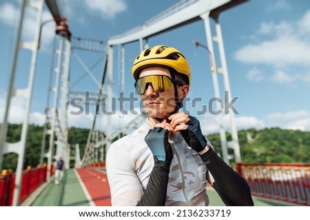 Portrait of a handsome male cyclist in a helmet and sporty outfit on the background of the bridge, looks away with a serious face and wears protection