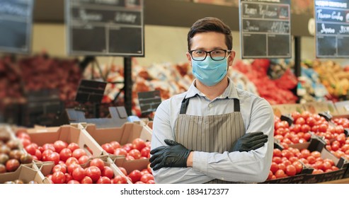 Portrait Of Handsome Male Food Store Worker In Medical Mask And Apron Standing In Grocery And Looking At Camera. Caucasian Man Seller Assistant Indoor With Vegetables On Background Supermarket Concept