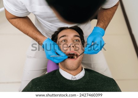 Portrait of handsome male patient lying in stomatology seat with open mouth during unrecognizable professional dentist examining teeth, with dental tool in dentistry clinic. Concept of stomatology. [[stock_photo]] © 