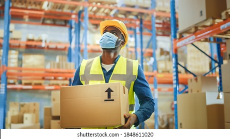 Portrait Of Handsome Male Worker Wearing Medical Face Mask And Hard Hat Carries Cardboard Box Walks Through Retail Warehouse Full Of Shelves With Goods. Safety First Protective Workplace.