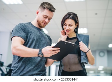 Portrait Of A Handsome Man And Attractive Woman Resting After Workout And Writing Something In Sports Gym. Personal Instructor With A Client Making A Training Plan