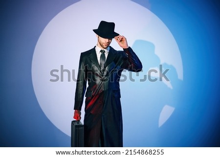 Portrait of handsome man in black coat and hat holding briefcase posing in the spotlight on studio background. noir film style. Private detective, spy, investigation concept. 