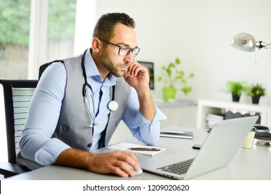 Portrait Of Handsome Man Male Doctor In Medical Practice Office Writing Prescription In Laptop Computer