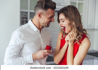 Portrait of a handsome man proposing to his happy girlfriend while holding engagement ring in a box and standing on a kitchen