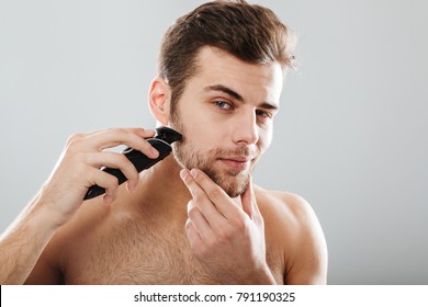 Portrait of handsome man shaving his beard with electric shaver in morning, against grey background