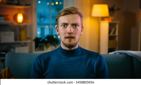 Portrait Of A Handsome Man Sitting On A Couch At Home At Night, Watching Gore Horror Movie On TV. He Gets Really Scared, Confused And Disgusted.