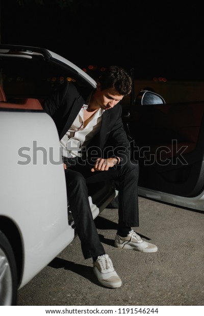 Portrait of a handsome man sitting posing in white\
cabriolet and looking on his watch. Nightlife. Businessman in suit\
in luxury car