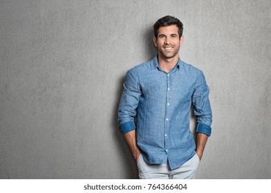 Portrait Of A Handsome Man Smiling Against Grey Background With Copy Space. Smiling Latin Guy With Hands In Pocket In Blue Shirt Standing And Leaning On Wall. Successful Hispanic Man Looking At Camera