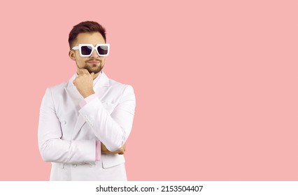 Portrait Handsome Man In Stylish Sunglasses Wearing In Classic White Suit And Pink Shirt, Keeps Hand Touching His Chin Standing Over Isolated Pink Background. Good Looking Guy Planning, Thinking