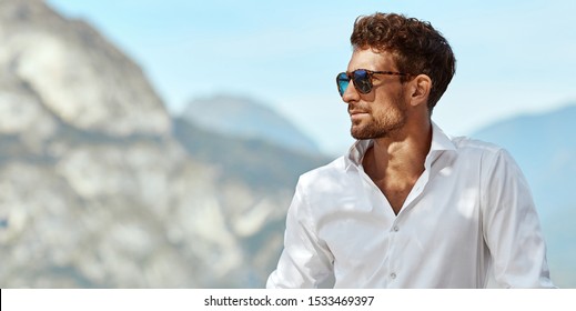 Portrait of handsome man wearing elegant white shirt and sunglasses, standing near the lake in the alps - Powered by Shutterstock