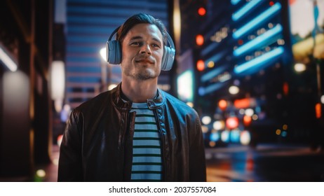 Portrait of Handsome Man Wearing Headphones Walking Through Night City Street Full of Neon Light. Smiling Stylish Man Listening to Music, Podcast, Talk Show. - Powered by Shutterstock