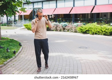 Portrait Of Handsome Mature Businessman Holding Messenger Bag, Wearing Stylish Eyeglasses Looking At Camera. Smiling Middle Aged Man Using Smartphone, Standing On The Street. Mobile Banking Concept