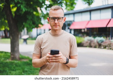 Portrait Of Handsome Mature Businessman Holding Messenger Bag, Wearing Stylish Eyeglasses Looking At Camera. Smiling Middle Aged Man Using Smartphone, Standing On The Street. Mobile Banking Concept