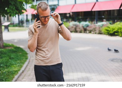 Portrait Of Handsome Mature Businessman Holding Messenger Bag, Wearing Stylish Eyeglasses Looking At Camera. Smiling Middle Aged Man Using Smartphone, Standing On The Street. Mobile Banking Concept