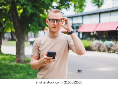 Portrait Of Handsome Mature Businessman Holding Messenger Bag, Wearing Stylish Eyeglasses Looking At Camera. Smiling Middle Aged Man Using Smartphone, Standing On The Street. Mobile Banking Concept