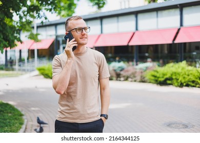 Portrait Of Handsome Mature Businessman Holding Messenger Bag, Wearing Stylish Eyeglasses Looking At Camera. Smiling Middle Aged Man Using Smartphone, Standing On The Street. Mobile Banking Concept