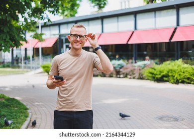 Portrait Of Handsome Mature Businessman Holding Messenger Bag, Wearing Stylish Eyeglasses Looking At Camera. Smiling Middle Aged Man Using Smartphone, Standing On The Street. Mobile Banking Concept