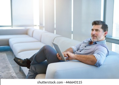 Portrait of a handsome mature businessman, relaxing on a couch in a business lounge, using a digital tablet and looking at the camera with a positive expression - Powered by Shutterstock