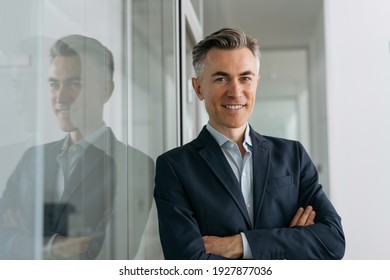 Portrait of handsome mature manager with arms crossed looking at camera, smiling standing in modern office 
