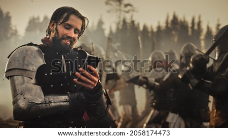 Portrait of Handsome Medieval Knight Using Smartphone on Battlefield, Smiling. Fun Concept: Successful Armored Warrior Ordering Online, Betting, Investing, doing E-commerce. War is Raging