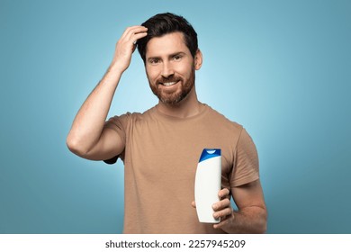 Portrait of handsome middle aged bearded man standing with shampoo bottle in hand, touching his hair and smiling, posing over blue background - Powered by Shutterstock