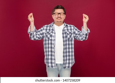 Portrait Of Handsome Middle Aged Man In Casual Checkered Shirt And Eyeglasses Standing With Raised Arms In Yoga Pose, Doing Meditating And Relaxing. Indoor Studio Shot, Isolated On Dark Red Background