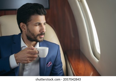 Portrait of handsome pensive middle eastern businessman drinking coffee, planning project looking at window sitting in airplane. Confident entrepreneur flying luxury private jet, successful business