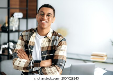 Portrait Of Handsome Positive Charismatic Hispanic Guy With Glasses, In Casual Shirt, Stand Near The Table With His Arms Crossed, Looks At Camera, Smiles Friendly. Male Designer, Freelancer Or Student