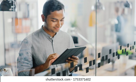 Portrait of Handsome Professional Indian Man Uses Touch Screen Digital Tablet Computer, Writes Important Email, Smiles Charmingly. Successful Man Working in Bright Diverse Office.