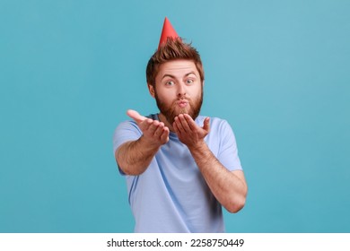Portrait of handsome romantic bearded man wearing party hat sending air kiss at camera, holiday greeting, flirt, celebrating birthday. Indoor studio shot isolated on blue background. - Shutterstock ID 2258750449