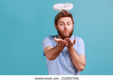 Portrait of handsome romantic young adult bearded angelic man with holy nimbus sending air kiss to camera, flirty angel, valentines day. Indoor studio shot isolated on blue background.