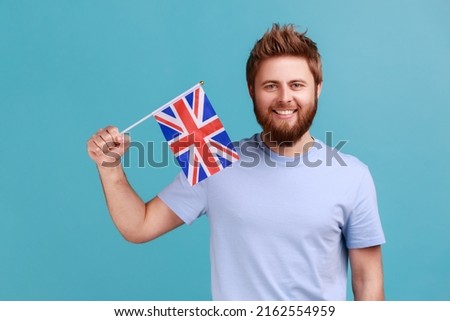 Portrait of handsome satisfied bearded man holding flag of a constituent unit of the United Kingdom, celebrating British Independence Day. Indoor studio shot isolated on blue background.