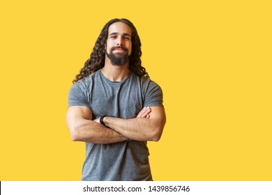 Portrait of handsome satisfied bearded young man with long curly hair in grey tshirt standing with crossed arms and looking at camera with smile. indoor studio shot isolated on yellow background.