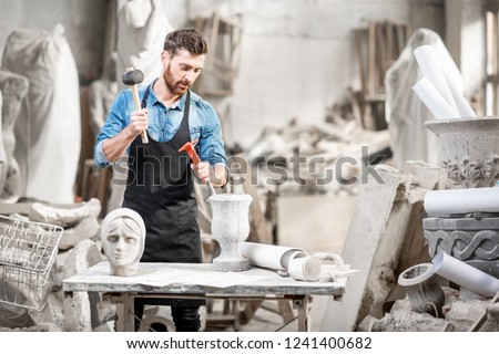 Portrait of a handsome sculptor in blue t-shirt and apron working with stone sculptures on the table at the old atmospheric studio