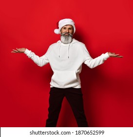 Portrait of a handsome senior bearded man, in a white hoodie and santa hat, stands on a red background with his hands spread apart