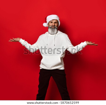 Portrait of a handsome senior bearded man, in a white hoodie and santa hat, stands on a red background with his hands spread apart