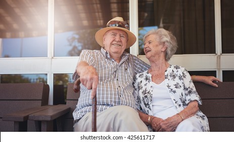 Portrait Of Handsome Senior Man Sitting With His Wife On A Bench Outside Their House. Retired Couple Taking A Break And Relaxing Outside Their Home.