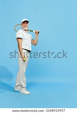 Portrait of handsome senior man in stylish white outfit posing with tennis racket over blue background. Concept of leisure activity, hobby, lifestyle, fitness, emotions, retro style