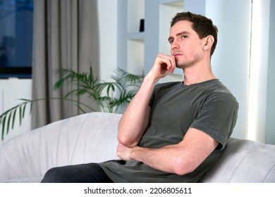 Portrait of handsome serious dreamy thoughtful pensive guy, young man sitting in living room on couch or sofa at home thinking, dreaming 