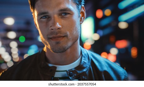 Portrait Of Handsome Serious Man Looking At Camera, Standing In Night City With Bokeh Neon Street Lights In Background. Confident Young Man With Stylish Hair. Close-up Shot