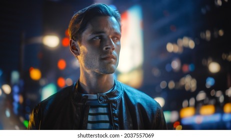 Portrait Of Handsome Serious Man Standing, Looking Around Night City With Bokeh Neon Street Lights In Background. Focused Confident Young Man Thinking. Portrait Shot.